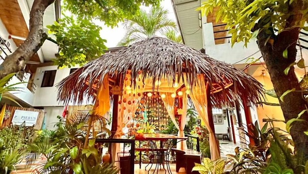A thatched-roof gazebo surrounded by lush greenery and potted plants, with a tiled floor and a visible sign for Pierus. - Papierus Pensionne (Kalibo)