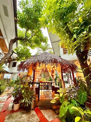 A thatched-roof gazebo surrounded by lush greenery and potted plants, with a tiled floor and a visible sign for Pierus. - Papierus Pensionne (Kalibo)