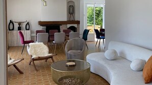 A modern living room with a curved white sofa, a round coffee table, and a fireplace. There are several chairs and a dining table in the background. - Villa Bien-être et Fête : Jacuzzi, Sauna, Court de Tennis, Piscine D'été, Étang (Cézy)