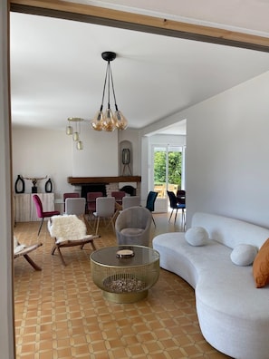 Una sala de estar moderna con un sofá blanco curvo, una mesa redonda de centro y una chimenea. Se ven varias sillas y una mesa de comedor al fondo.