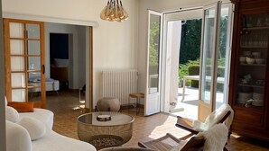 A spacious living room with a white sofa, a round coffee table, and a white armchair. There is a glass door leading to a balcony with a view of greenery. - Villa Bien-être et Fête : Jacuzzi, Sauna, Court de Tennis, Piscine D'été, Étang (Cézy)