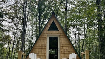 Situated in a forested area, this cabin features a front door and a cozy porch with two Adirondack chairs.