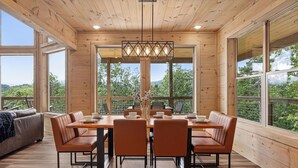A wooden dining area with a large table and chairs, large windows, and a view of greenery.