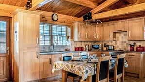 Une cuisine aménagée dans le style d’un chalet en bois, avec une table à manger pour quatre personnes, une horloge et une fenêtre équipée de stores.