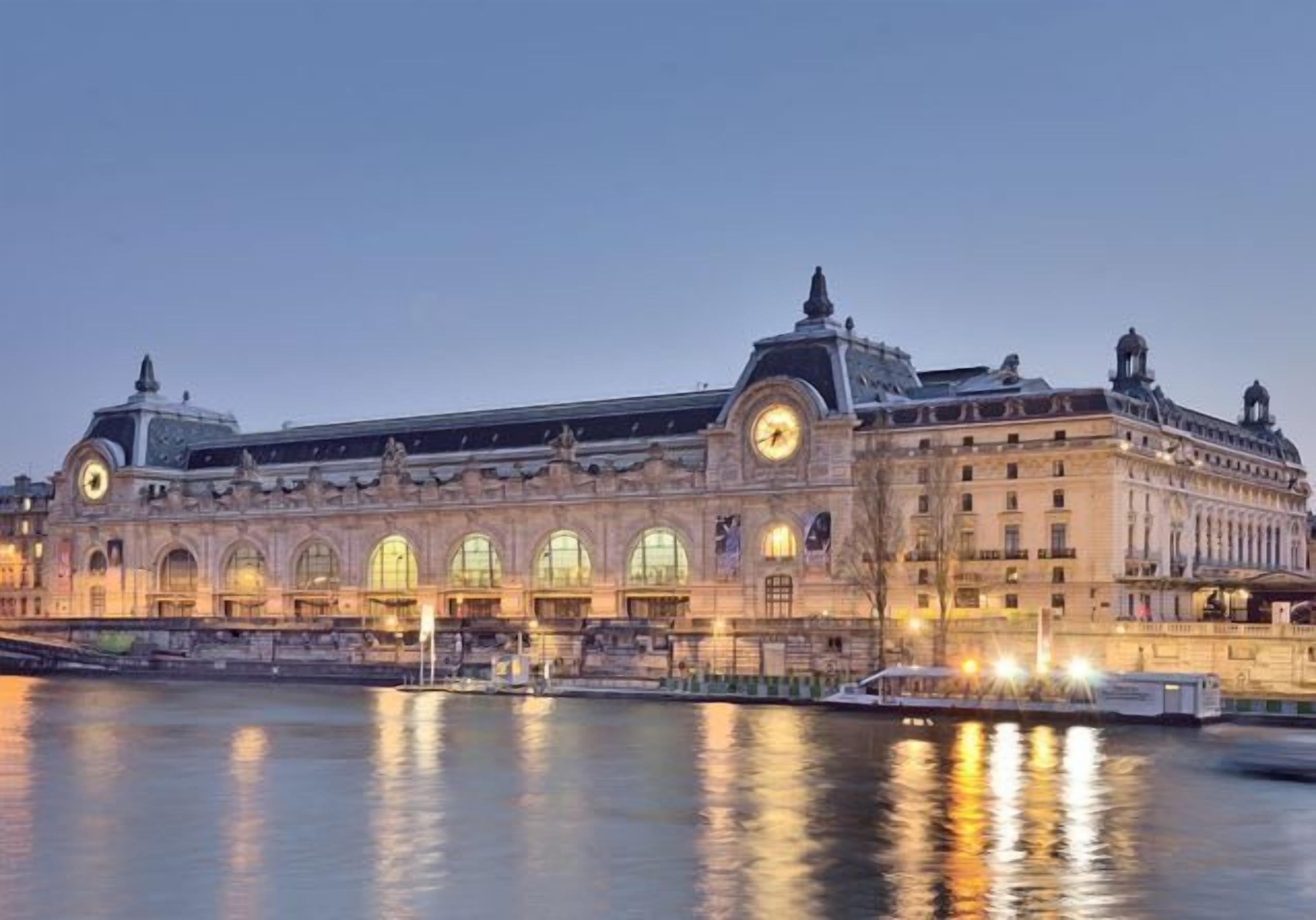 Un grand bâtiment historique doté d’arcs en plein cintre et surmonté d’une tour horloge, situé au bord d’un plan d’eau.