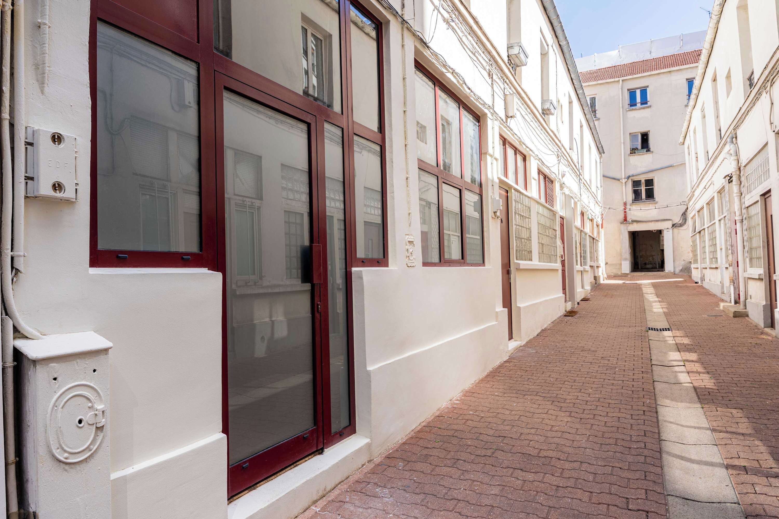 A narrow alleyway with white buildings, red-framed windows, and a brick pathway.