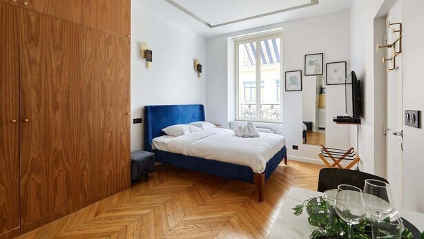 A bedroom with a large wardrobe, a bed with white bedding, a herringbone-patterned wooden floor, and a window with a view of the outside. - Cosi apartment Bastille-Le Marais (Paris)