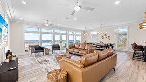 A spacious living room with a large tan sofa, a dining area with a table and chairs, and a view of the ocean through large windows. - Water's Edge: Brand New Luxury Beachfront Home with Elevated Pool. Pet Friendly1 (Mexico Beach)