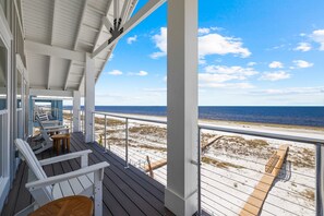 Eine überdachte Terrasse mit weißen Stühlen und einem Holztisch, mit Blick auf einen Sandstrand und das Meer.