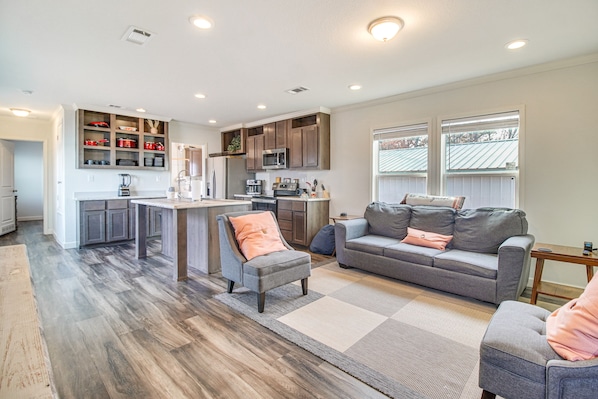 A modern kitchen with built-in appliances, a dining area, and a living room with a grey sofa and ottoman.