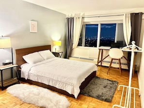 A hotel room with a bed, bedside table, lamp, window with curtains, and a view of the city at night.