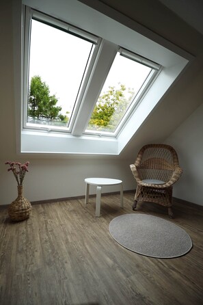 Ein Zimmer mit geneigter Decke, zwei Oberlichtern, einem Korbstuhl, einem kleinen runden Tisch und einem gewebten Korb.