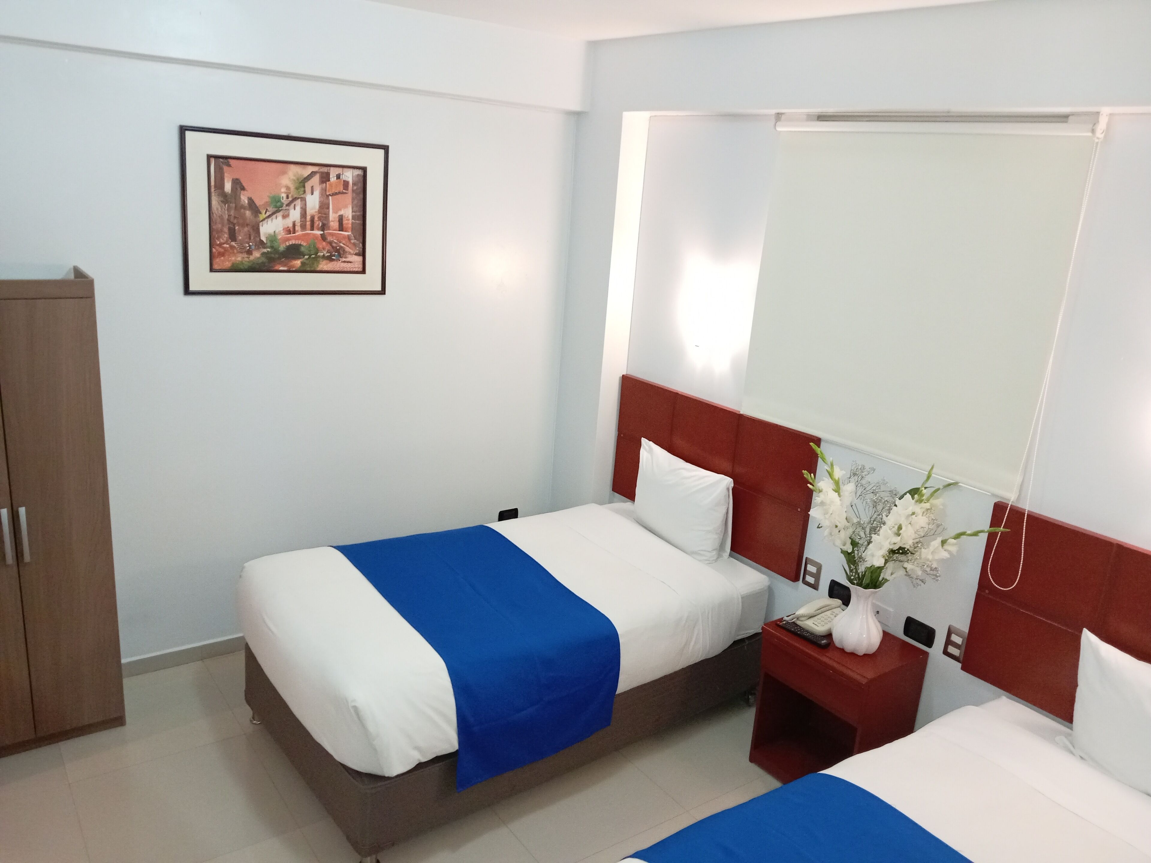 A hotel room with two beds, a wooden wardrobe, a framed picture on the wall, a bedside table with a phone and a vase of flowers, and a window with a white blind.