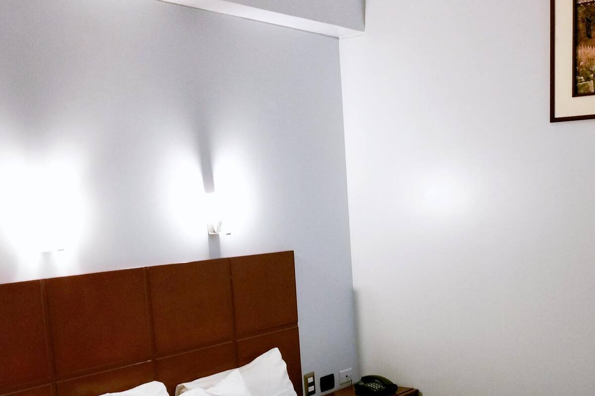 Ein ordentlich bezogenes Bett mit weißen Leinen und einer roten Tagesdecke, ein hölzerner Kopfteil, ein Nachttisch mit Telefon und ein gerahmtes Bild an der Wand.