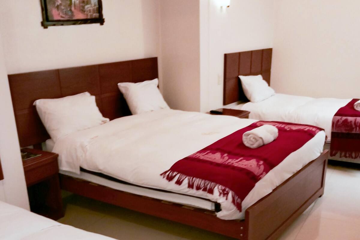 Ein Hotelzimmer mit zwei Betten, jeweils mit einem hölzernen Kopfteil, weißer Bettwäsche und einer roten Decke. Ein Nachttisch mit Lampe und ein gerahmtes Bild an der Wand sind ebenfalls vorhanden.