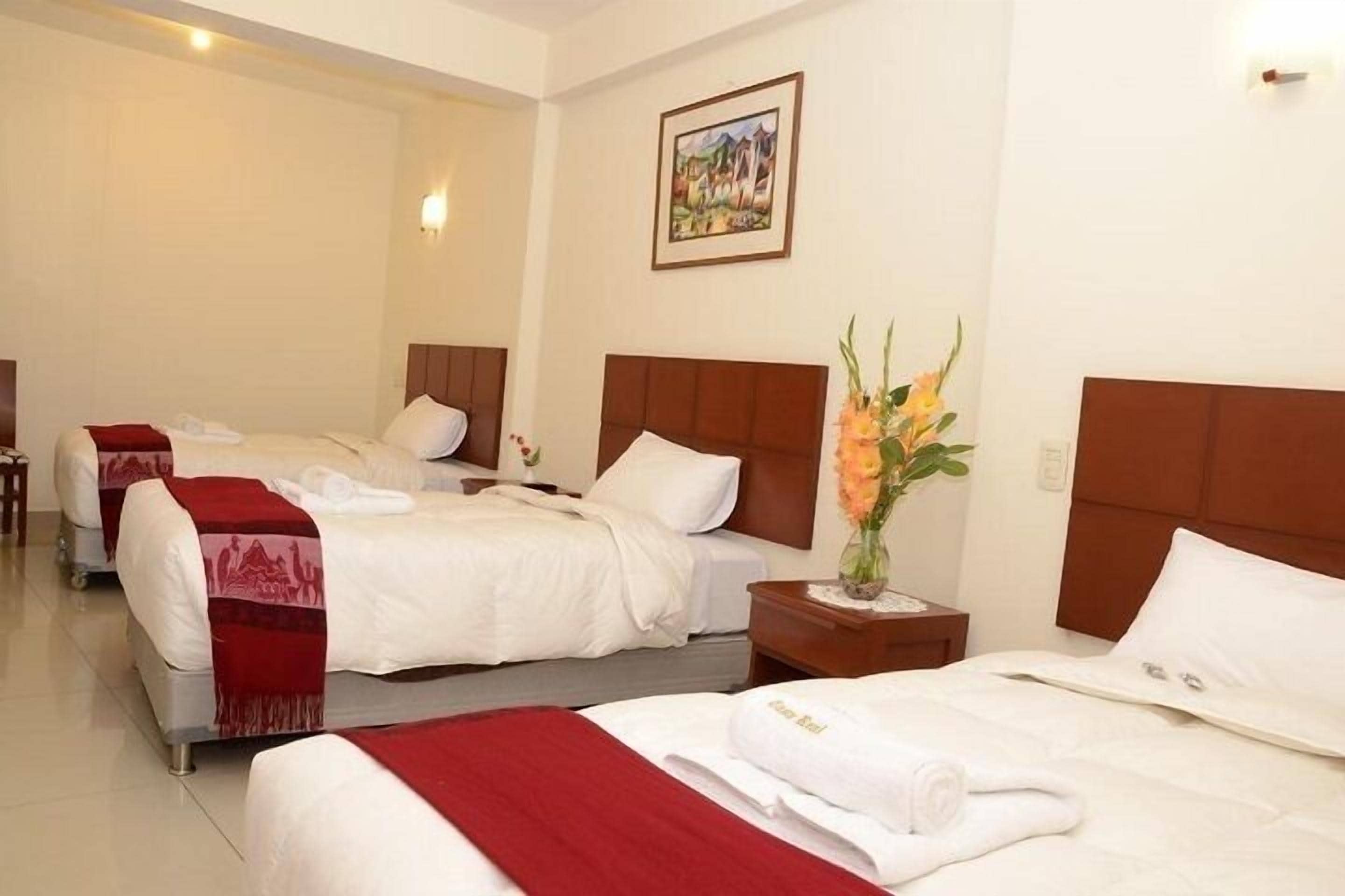 A hotel room with three beds, each with white bedding and red throw blankets. There is a painting on the wall and a vase with flowers on the nightstand.