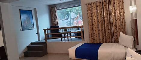 A hotel room with a bed, a chair, a small table with a plant, a window with curtains, and a staircase.