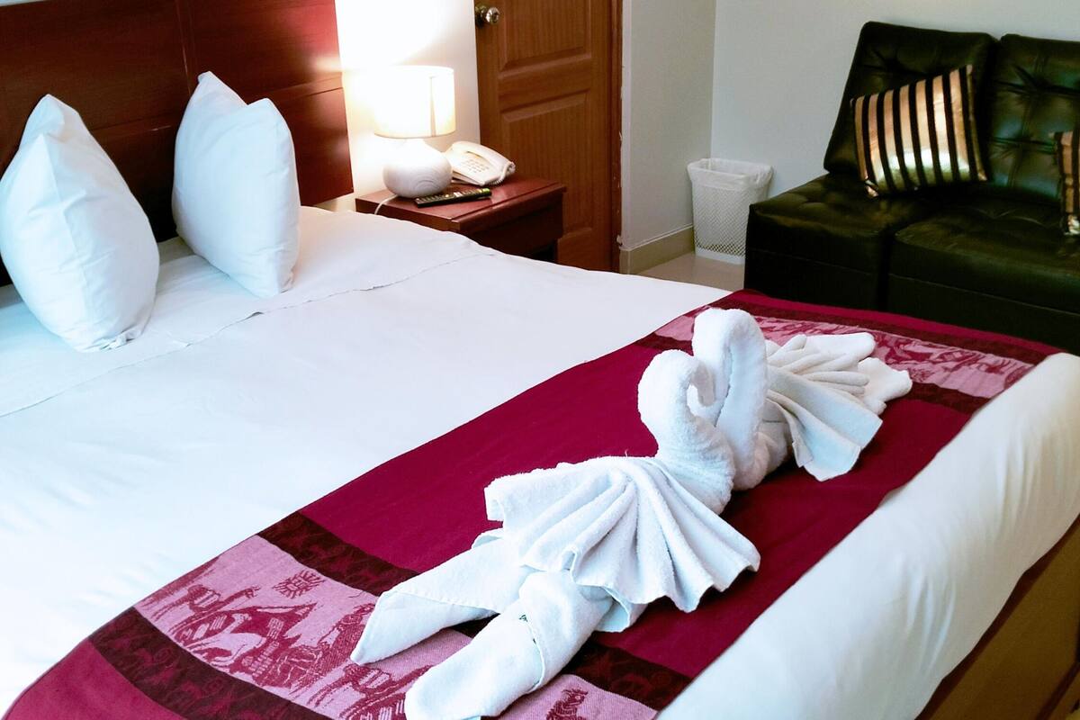 Ein Hotelzimmer mit einem ordentlich bezogenen Bett, einem Holzkopfteil, einem Nachttisch mit Lampe und Telefon sowie einem grünen Sofa mit gestreiften Kissen.