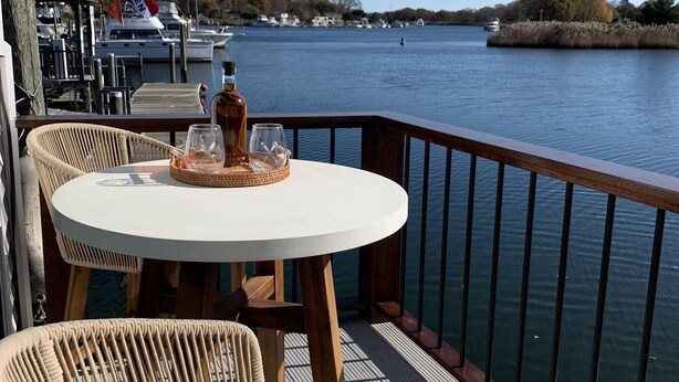 Une table ronde entourée de chaises en osier, placée sur une terrasse surplombant une étendue d’eau calme, avec des bateaux et un quai en arrière-plan.
