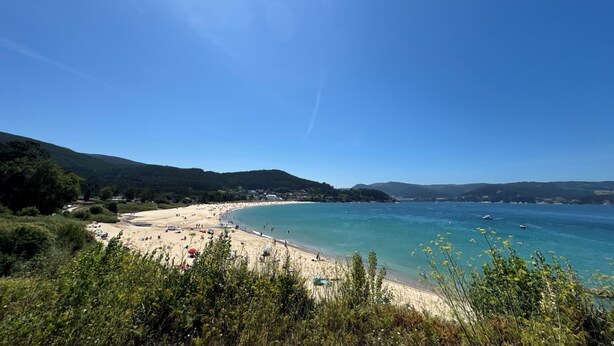 Una playa con agua azul cristalina, orilla de arena y colinas verdes al fondo.