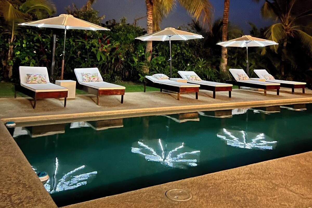 Un área junto a la piscina con sillones de descanso y sombrillas, rodeada de palmeras.