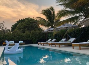 A swimming pool with lounge chairs and umbrellas, surrounded by palm trees and greenery. - Arganama (San Jose)