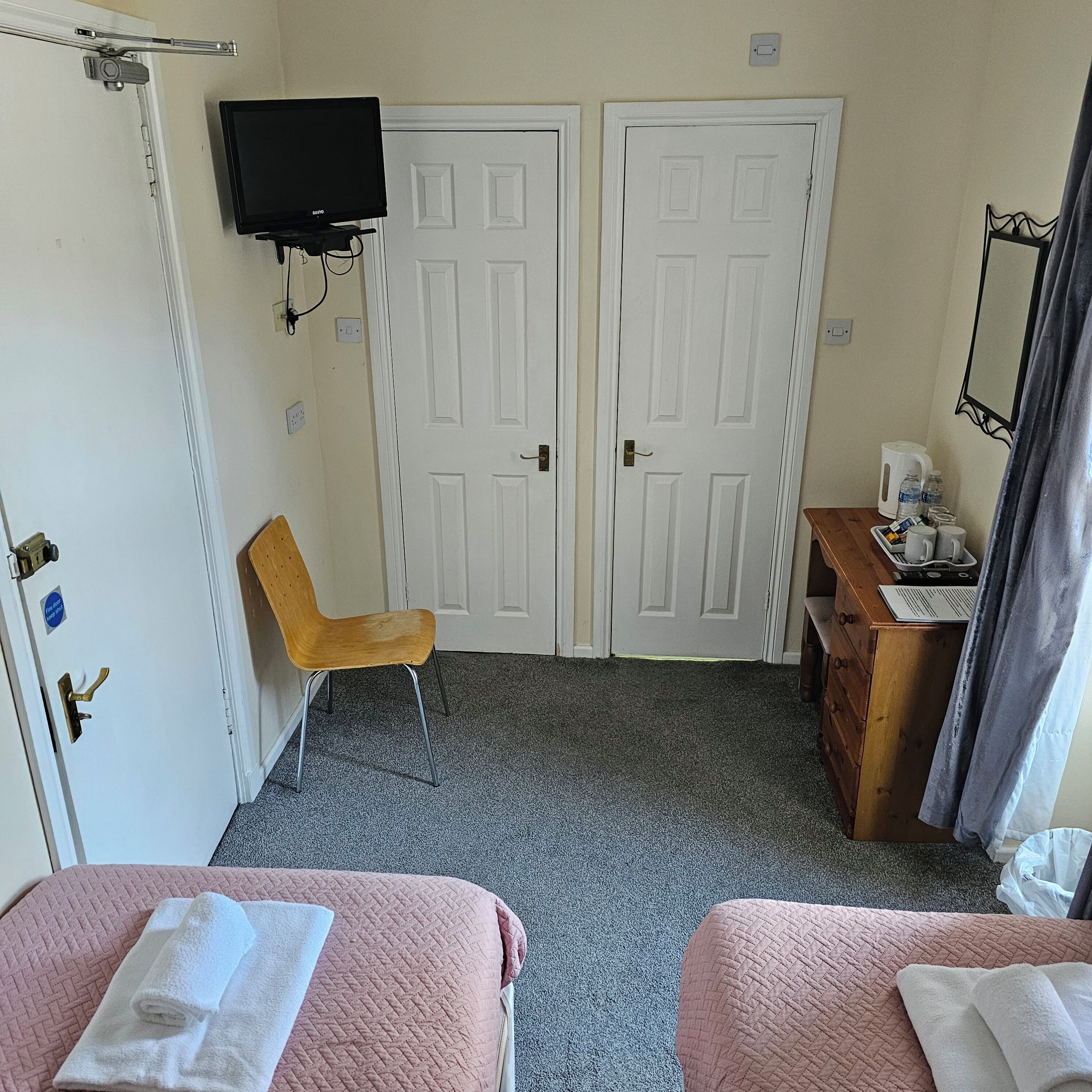 Een hotelkamer met twee bedden, een aan de muur bevestigde televisie, een houten stoel, een houten dressoir en een bureau met waterkoker en bekers.
