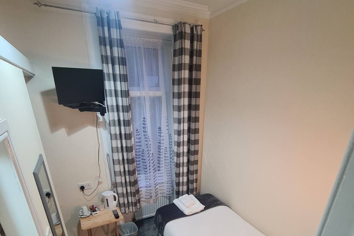 Pieni, siististi järjestetty makuuhuone, jossa on yksi sänky, työpöytä, jossa on vedenkeitin, roskakori ja seinälle asennettu televisio.