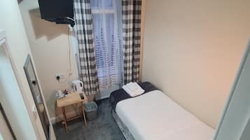 A small, neatly arranged bedroom with a single bed, a desk with a kettle, a trash bin, and a TV mounted on the wall.