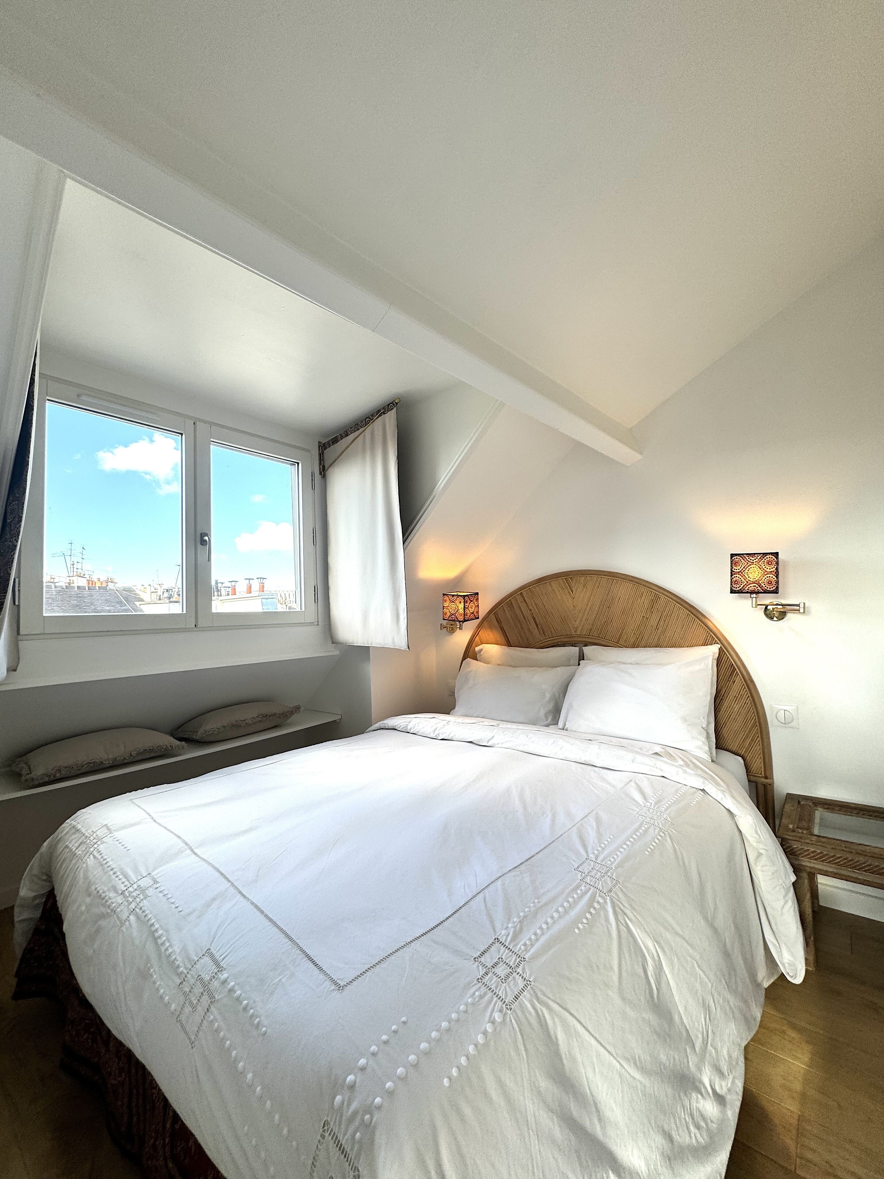 Un dormitorio con una cama grande, una ventana con cortinas, una lámpara de pared y vistas de la ciudad.