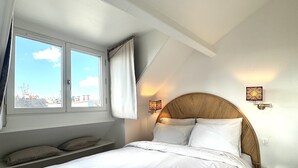 Ein Schlafzimmer mit einem großen Bett, einem Fenster mit Vorhängen, einer Wandleuchte und Blick auf die Stadt.