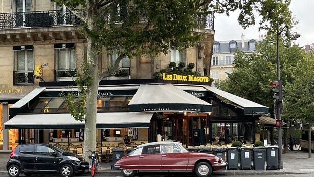 Straßenansicht mit einem Vintage-Roten Auto, das an einer Ecke geparkt ist, einem schwarzen Auto und einem Gebäude mit dem Schild "Les Deux Magots" Café.