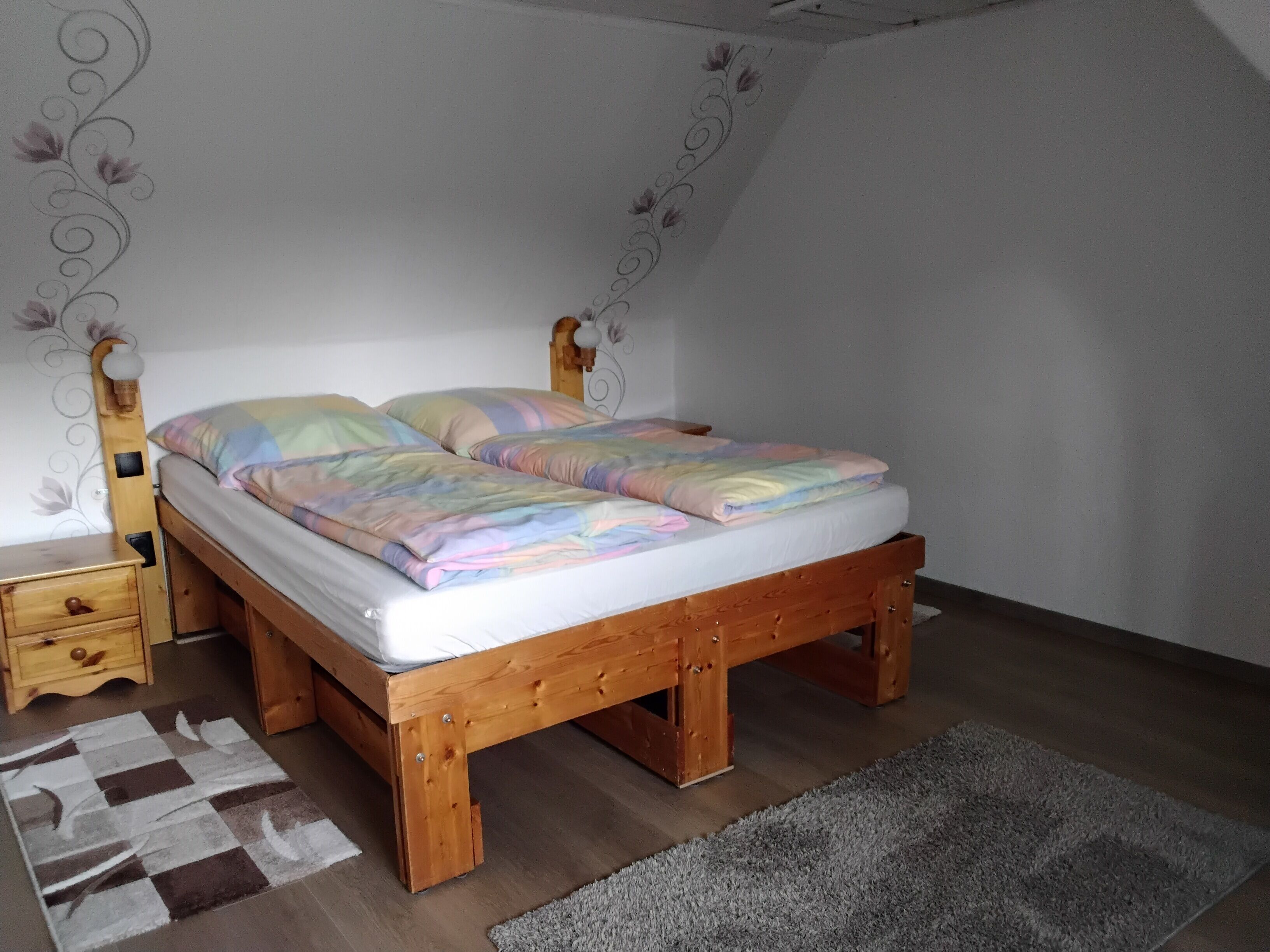 Un lit simple avec un cadre en bois, une table de chevet, une commode et un tapis à motifs dans une pièce aux murs blancs et au plafond en pente.