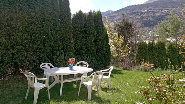 Mesa blanca al aire libre con cuatro sillas, rodeada de césped verde y árboles, con vistas a un paisaje montañoso.
