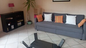 Una sala de estar con un sofá gris, una mesa de centro de vidrio, una planta en maceta y una estantería.