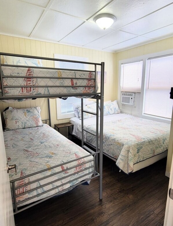 A dormitory room with bunk beds, a bedside table, and a window with blinds.