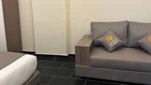 Ein modernes Hotelzimmer mit grauem Sofa, zwei passenden Kissen, einem Holznachttisch und einem Bett mit grauem Kopfteil.