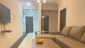 Ein modernes Wohnzimmer mit einer Couch, einem Couchtisch mit Obst, einem an der Wand befestigten Fernseher und einer Kristallleuchtre.