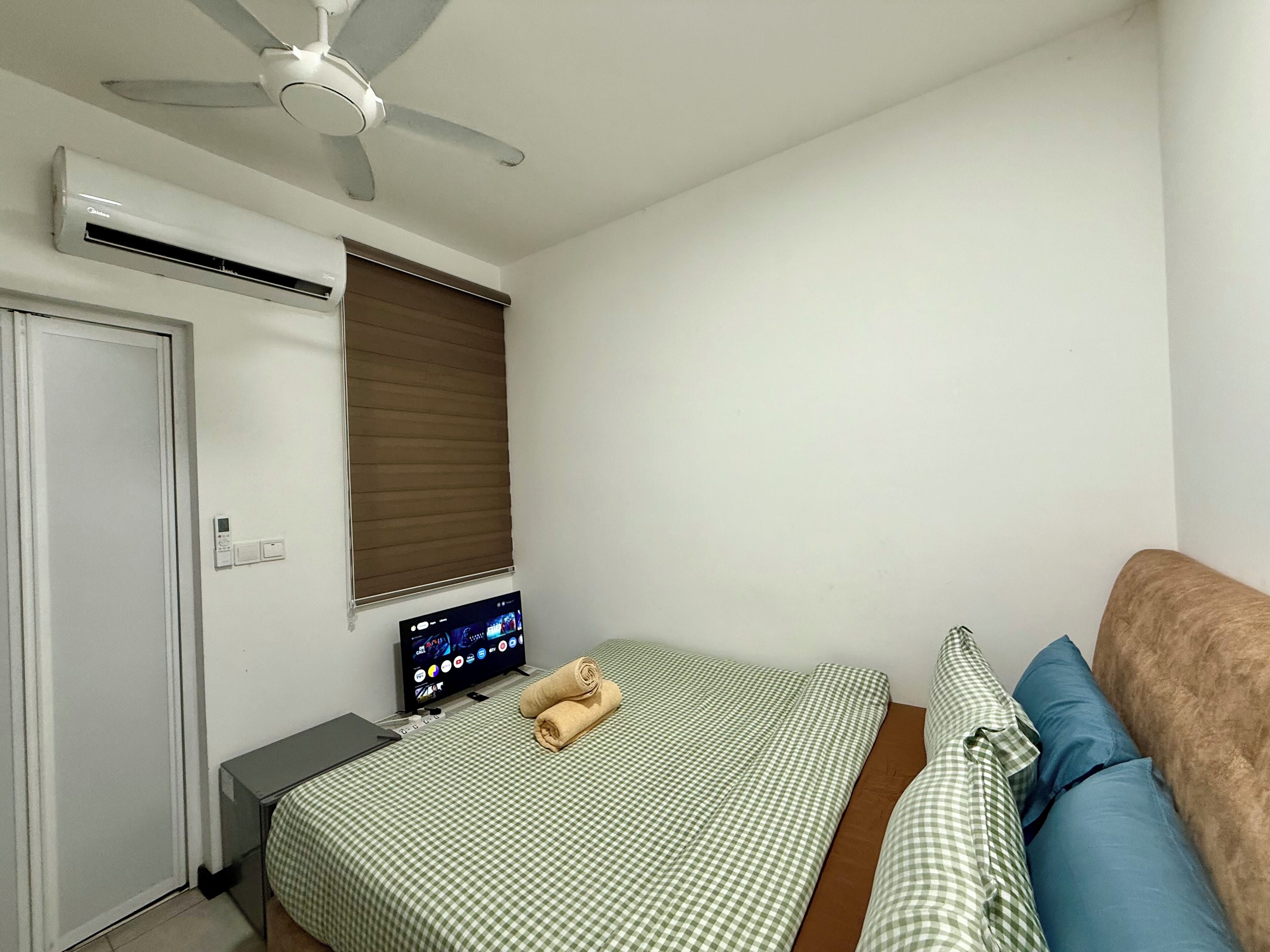 A bedroom with a bed, a television, an air conditioning unit, a ceiling fan, a window with blinds, and a door.