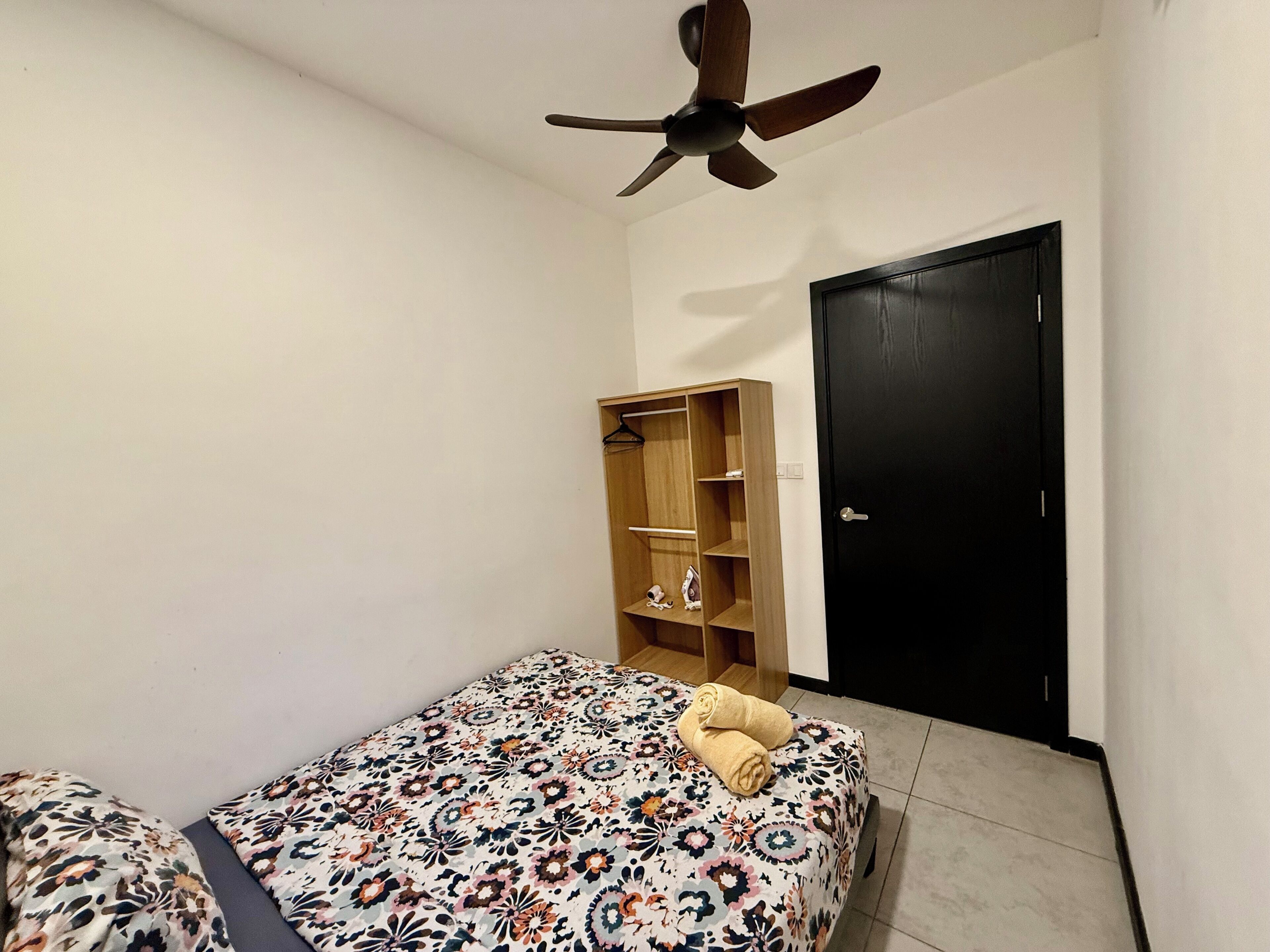 A small, clean room with a bed, a wooden wardrobe, a ceiling fan, and a black door.