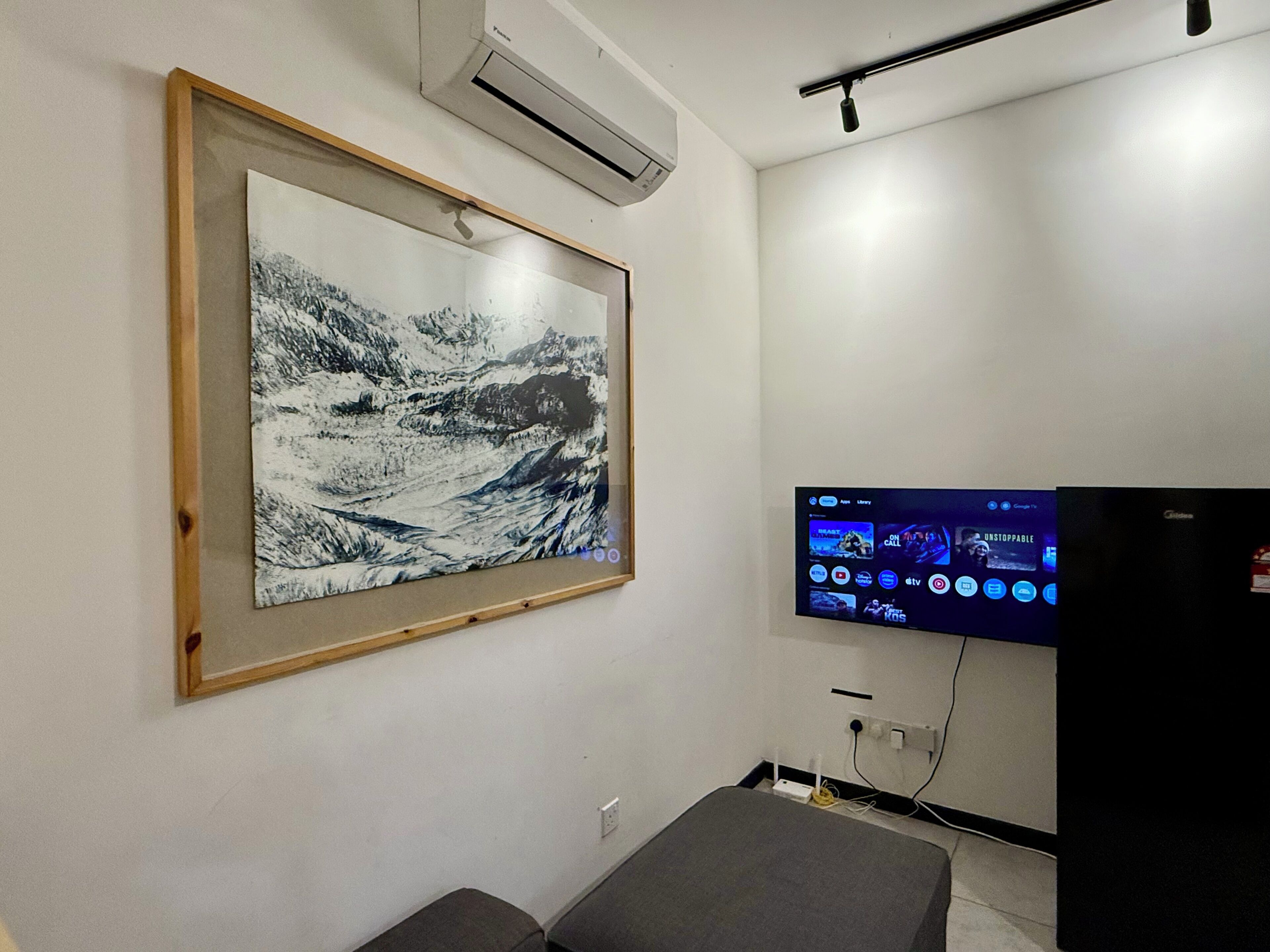 Ein Zimmer mit einem gerahmten Schwarz-Weiß-Berglandschaftsbild, einem montierten Flachbildfernseher, der verschiedene Sender zeigt, und einer kleinen grauen Bank.