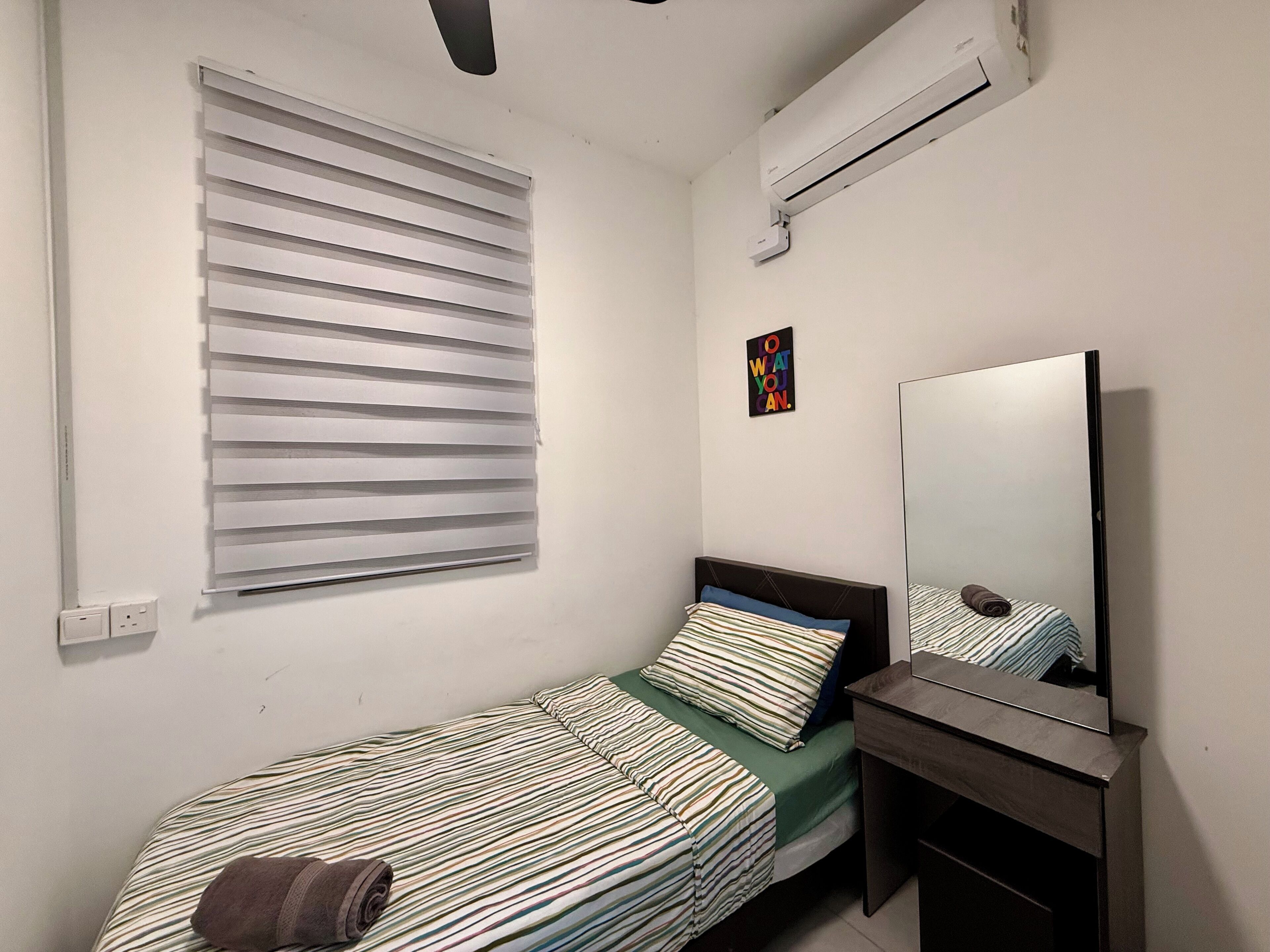 A small, neatly arranged bedroom with a single bed, a bedside table, a mirror, and a window with blinds.