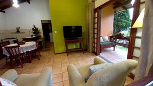A living room with a television, a dining area with chairs and a table, and a balcony with a view of greenery.