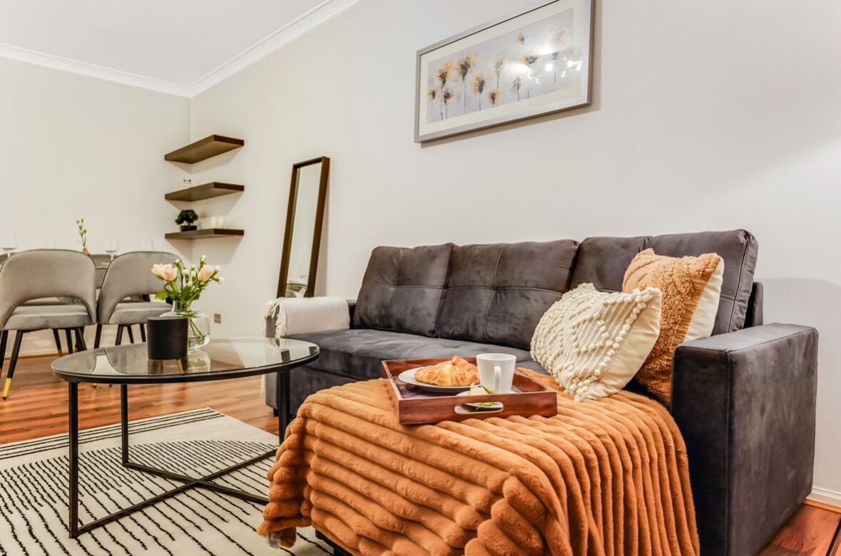 Ein modernes Wohnzimmer mit einem dunkelgrauen Sofa, einem Glastisch und einem braunen Teppich. Zwei graue Sessel, ein kleiner Beistelltisch mit einer Vase und ein gerahmtes Bild an der Wand ergänzen den Raum.