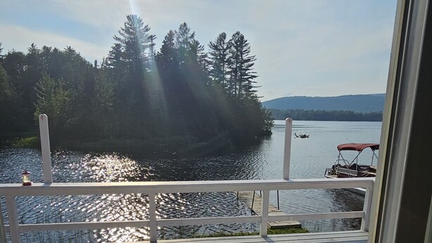 A wooden deck with a railing, overlooking a calm body of water, a boat, and a forested area. - Cozy Lake Front home for Boating, fishing, winter sports, and gorgeous views,  (Errol)