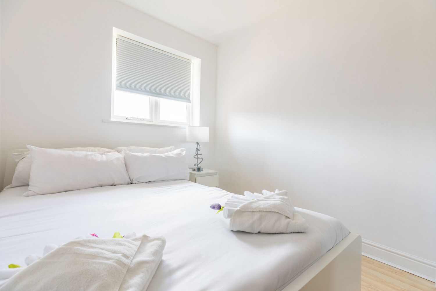 A minimalist bedroom with a large bed, white bedding, a bedside table with a lamp, and a window with blinds.