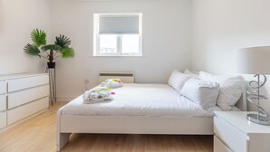A minimalist bedroom with a bed, a window, a radiator, a bedside table, and a potted plant. - Skyline Haven Modern 2BR w/ Free Parking & 4kTV (London)