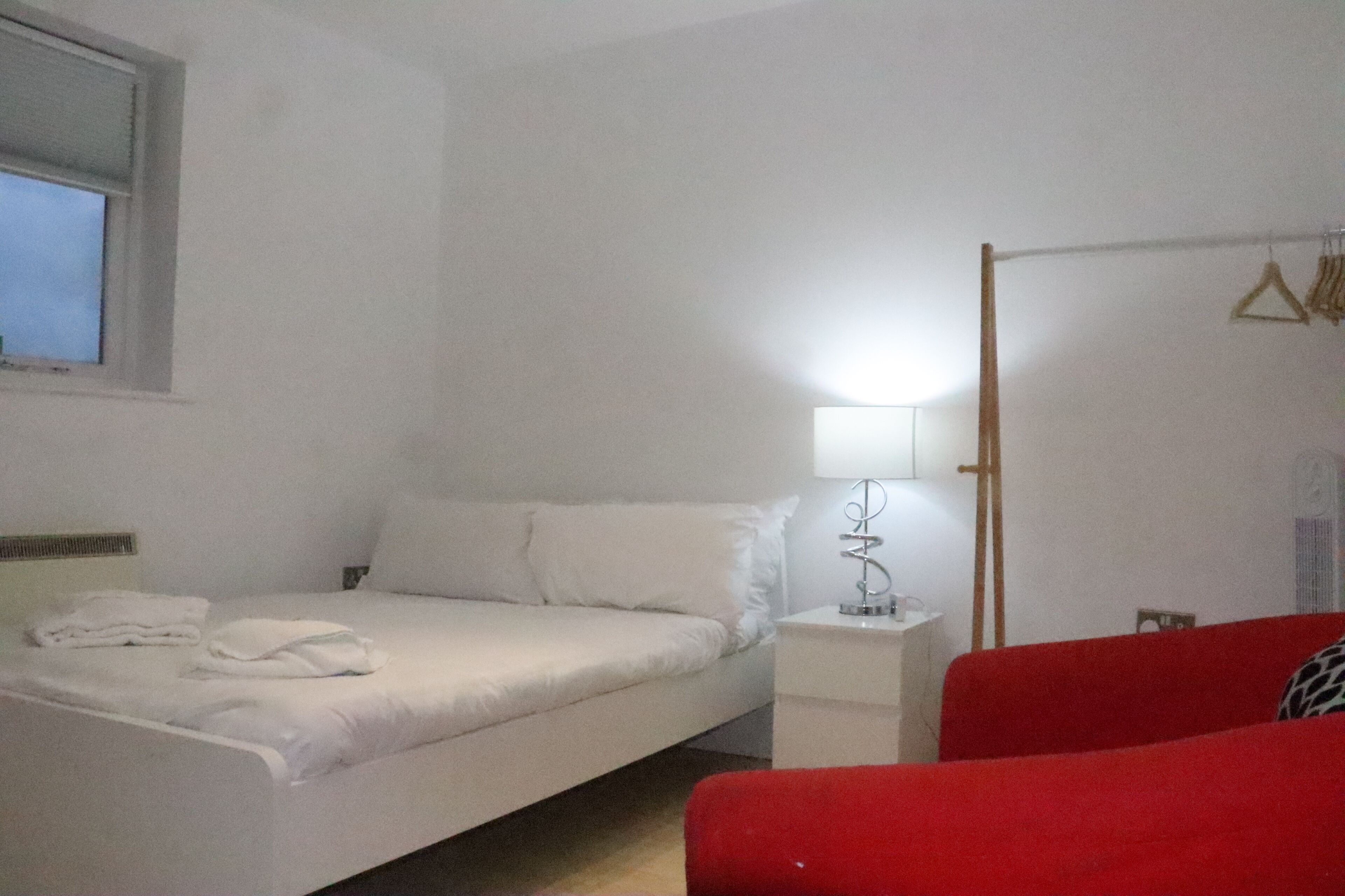 Un dormitorio con cama, mesita de noche con lámpara, una silla roja y un perchero.