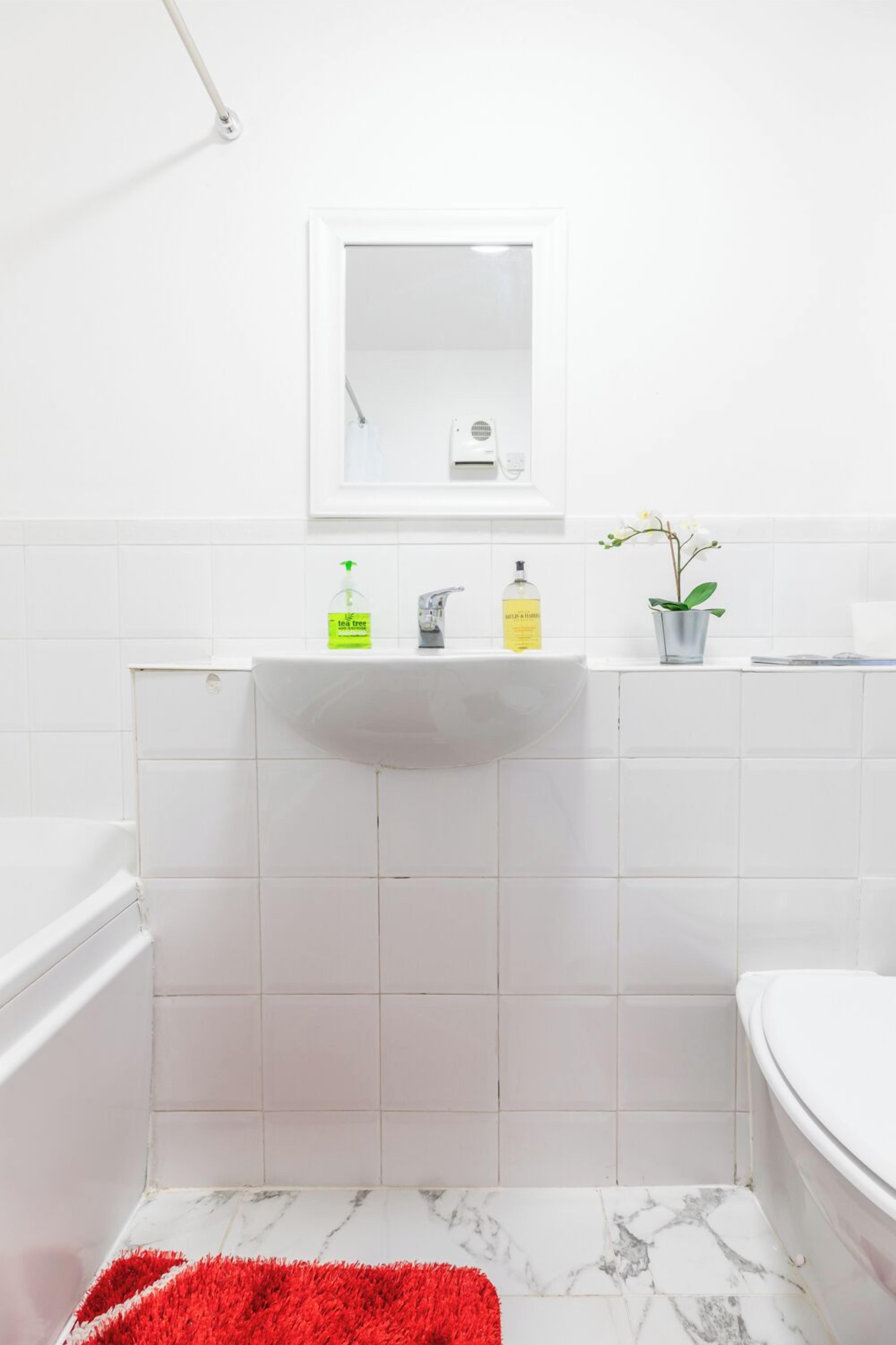 Un ba ño con lavamanos blanco, espejo e inodoro. Hay artículos de aseo en el lavamanos y una planta en maceta sobre la encimera.