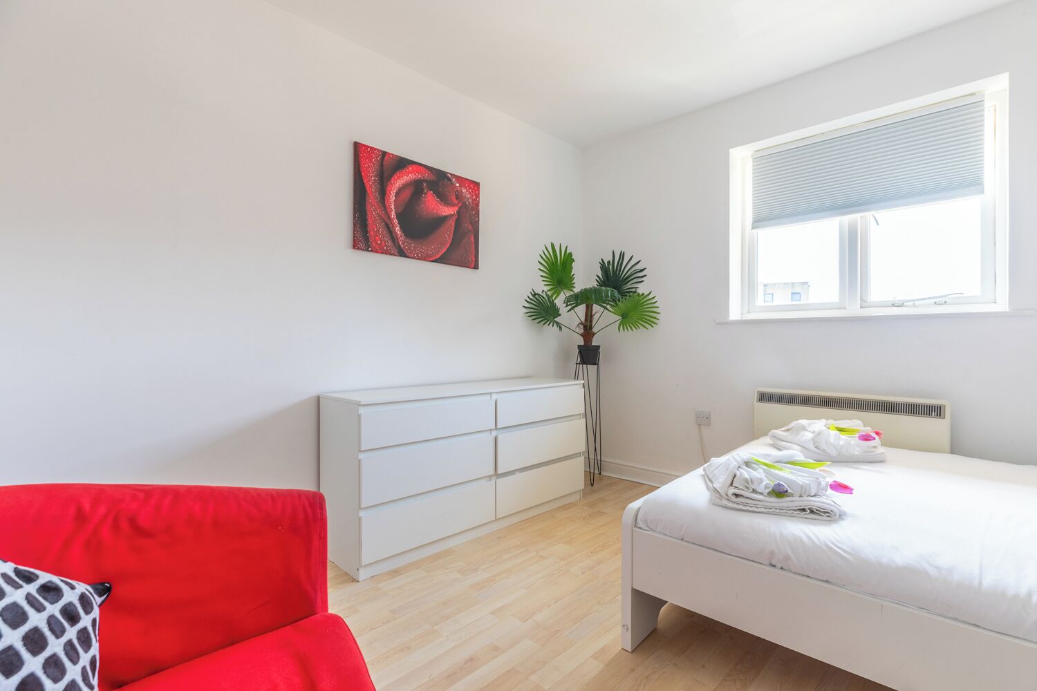 Une chambre moderne avec un lit, un canapé rouge, une commode blanche, une plante en pot et une fenêtre avec des stores.